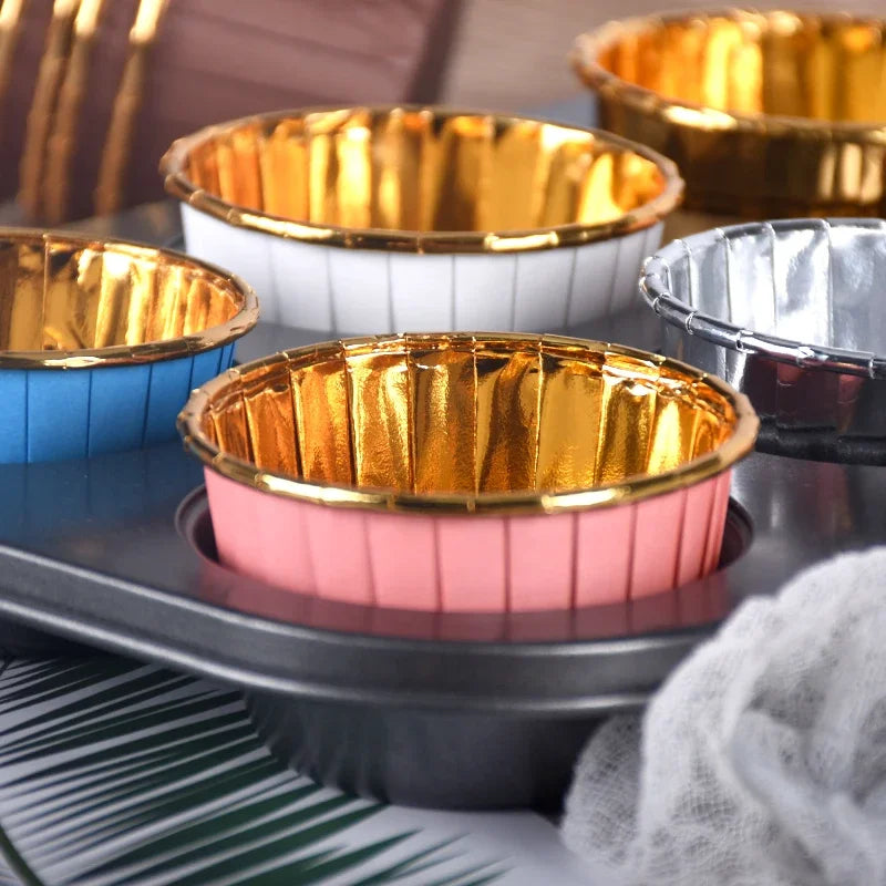A collection of golden greaseproof cupcake liners in various colors and finishes displayed on a dark tray.