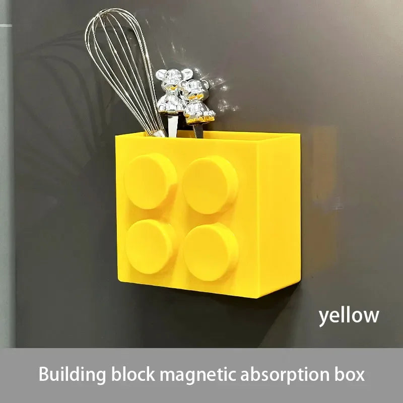Yellow building block magnetic absorption box with silver whisk and gold-colored charms mounted on a gray wall.