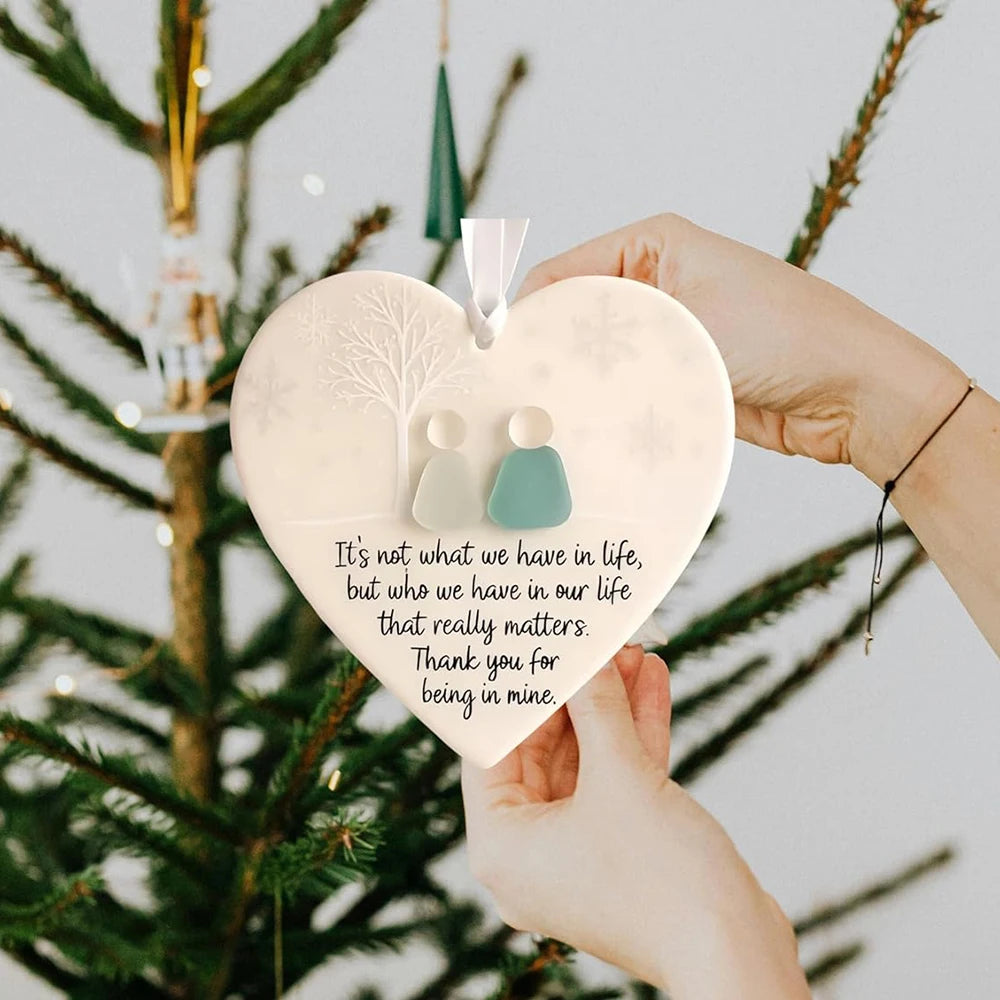 A hand holds a heart-shaped sea glass ornament with a keepsake pendant against a green Christmas tree backdrop.