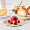 A slice of cake with cream is served on a disposable paper plate with fresh strawberries and sauce.