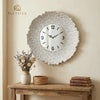 A round silver sunburst wall clock with a white face hangs on a beige wall above a wooden shelf.