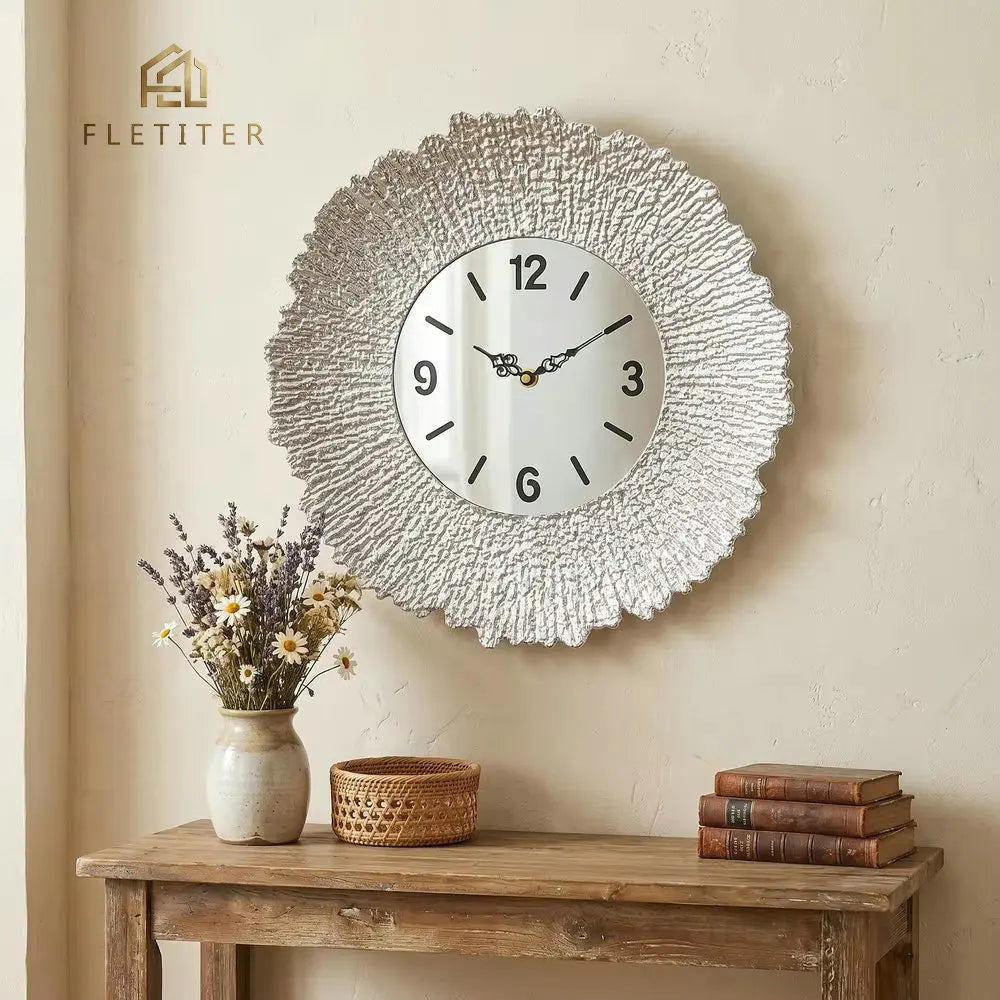 A round silver sunburst wall clock with a white face hangs on a beige wall above a wooden shelf.