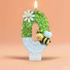 A large number zero decorated with green glitter, a white flower, and a bee figurine, set against a soft orange background.