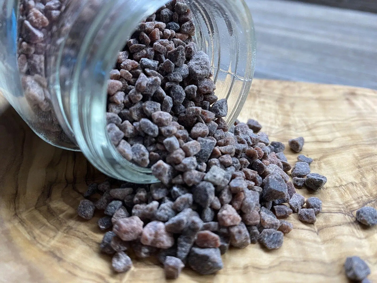 A glass jar filled with dark black salt is tipped over onto a wooden surface.