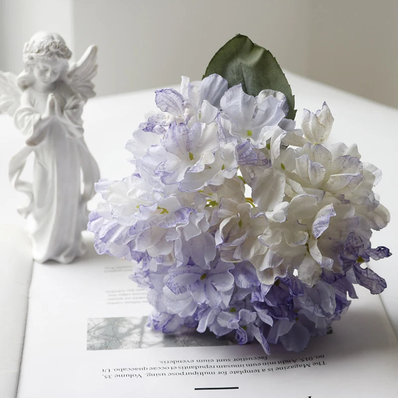 A bouquet of white and purple silk hydrangea flowers next to a white figurine.