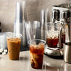 A close-up of several iced coffee drinks in clear plastic cups and a metal coffee dispenser.