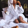 Eine Frau in einem weißen Bademantel entspannt sich im Bett, hält Essen und wirkt nachdenklich.