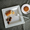 A white square plate holds a piece of bread, some nuts, and several dark chocolate truffles, next to a cup of coffee.