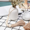 A handmade wood and jute bead garland displayed next to a white ceramic vase with dried flowers on a black grid surface.