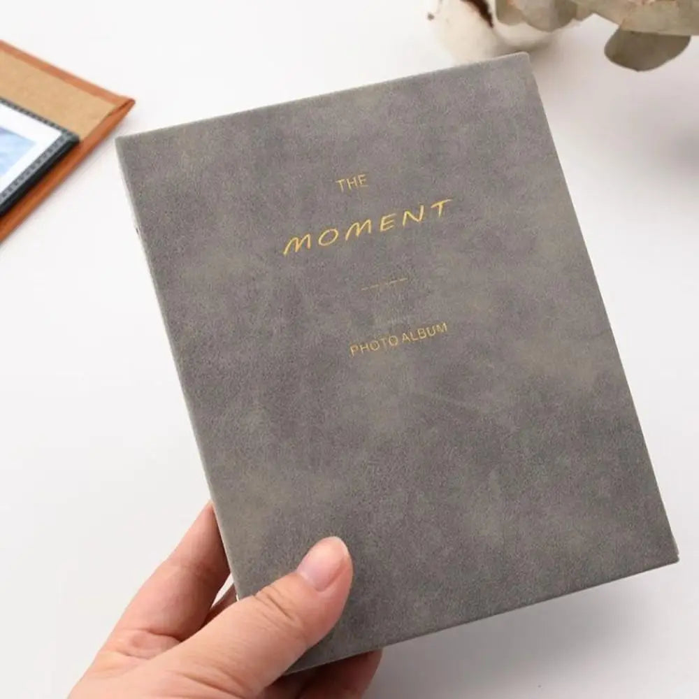 A hand holds a gray photo album titled The Moment Photo Album with gold lettering on a white surface.