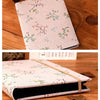 A light pink linen photo album with delicate green and pink floral embroidery resting on a wooden surface.