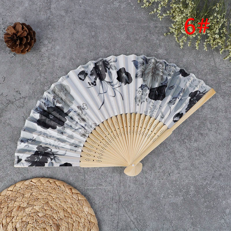 A hand carved bamboo folding fan with a gray and black floral pattern is displayed on a gray surface next to a pine cone and 