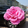 A large, vibrant pink artificial peony flower is displayed on the hood of a dark car.