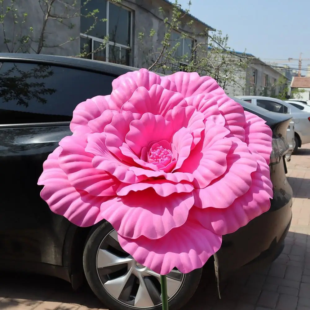 A large, vibrant pink artificial peony flower is displayed on the hood of a dark car.