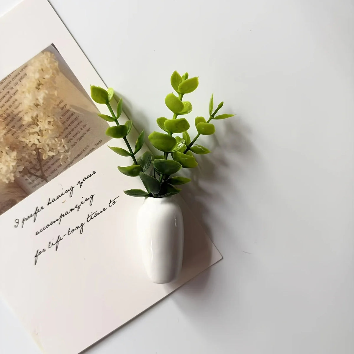 A white ceramic vase holds a sprig of bright green artificial flowers on a white surface.