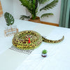 A realistic, olive-green and yellow patterned snake plush toy is displayed on a white surface next to potted plants.