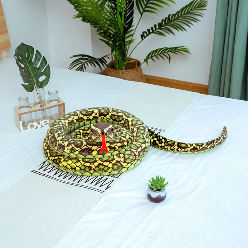 A realistic, olive-green and yellow patterned snake plush toy is displayed on a white surface next to potted plants.