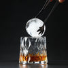 A hand holds a clear ice sphere above an Old Fashioned whiskey glass filled with amber liquid.