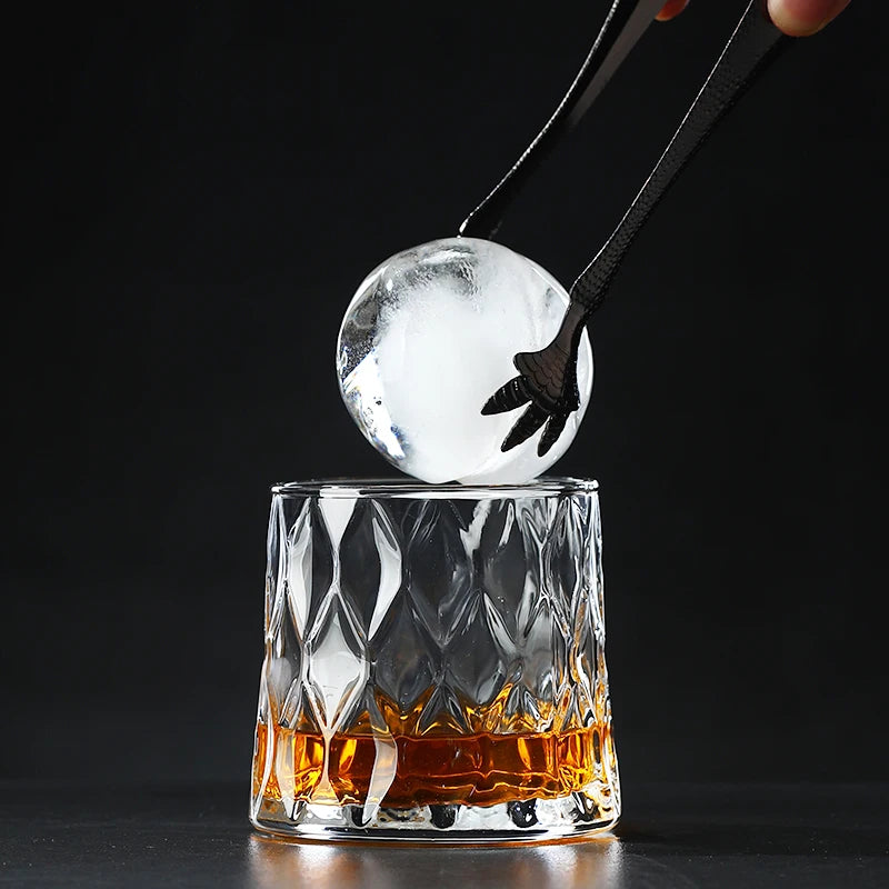 A hand holds a clear ice sphere above an Old Fashioned whiskey glass filled with amber liquid.