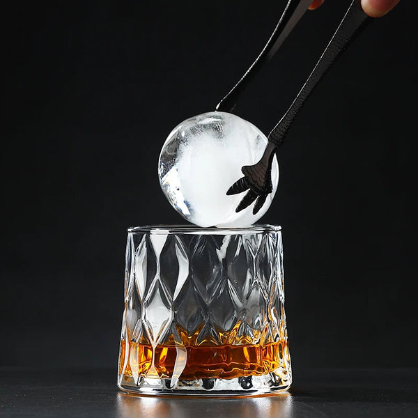 A hand holds a clear ice sphere above an Old Fashioned whiskey glass filled with amber liquid.