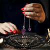 A hand with red nail polish holds a silver pendant on a wooden pendulum board for divination.
