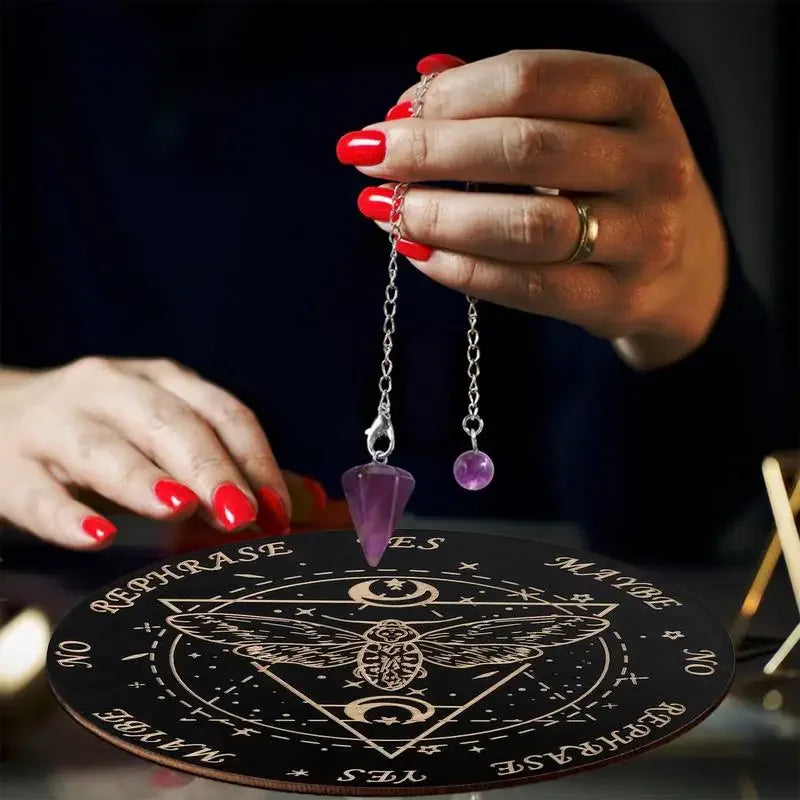 A hand with red nail polish holds a silver pendant on a wooden pendulum board for divination.