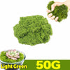 A hand holds a clump of bright green artificial moss next to a pile of the same moss and a potted succulent.