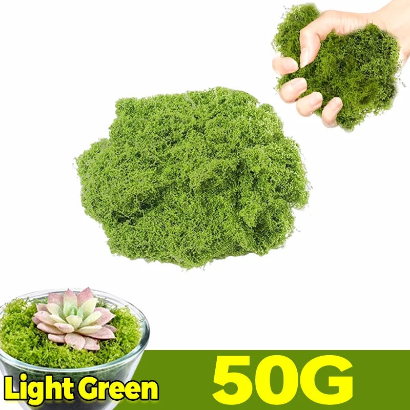 A hand holds a clump of bright green artificial moss next to a pile of the same moss and a potted succulent.