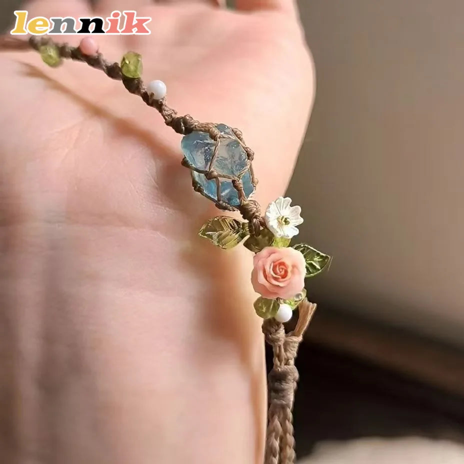 A macrame bracelet featuring a raw aquamarine crystal and a small pink rose accent on a plant stem.