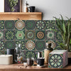 A close-up of self-adhesive tile wall stickers featuring intricate green and beige floral patterns on a wooden surface.