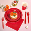 A red paper party tableware set featuring dinner plates, napkins, cups, and cutlery arranged on a pink surface.