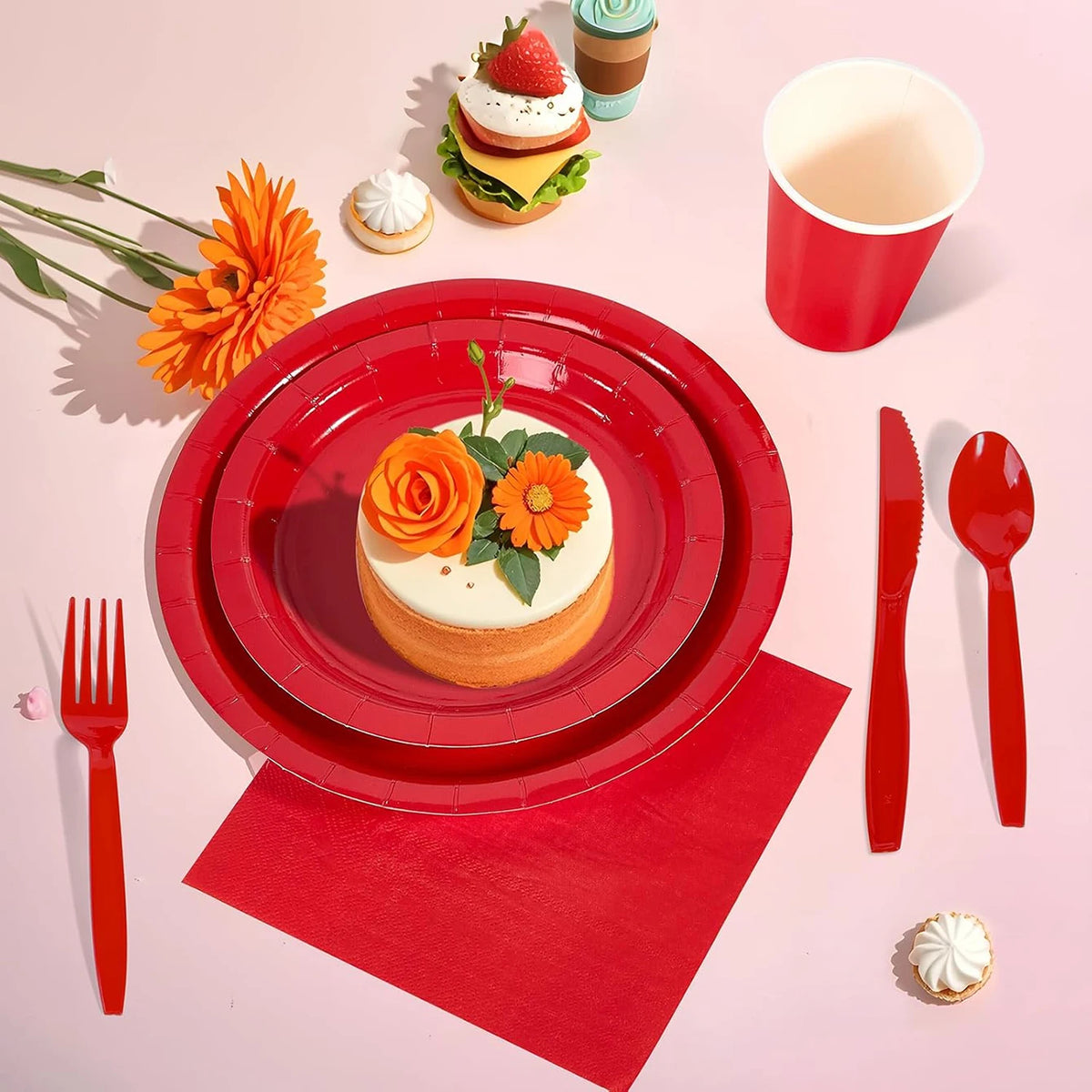A red paper party tableware set featuring dinner plates, napkins, cups, and cutlery arranged on a pink surface.