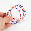 A hand holds a circular beaded stretch bracelet featuring red, white, and blue beads and gold stars.