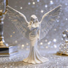 A white figurine of a praying angel with large wings is displayed on a sparkling surface.