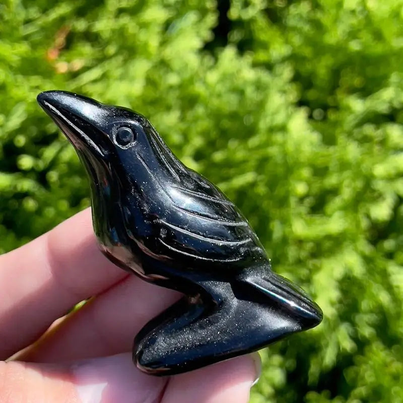 A handcrafted black obsidian crow statue held in a hand against a blurred green background.