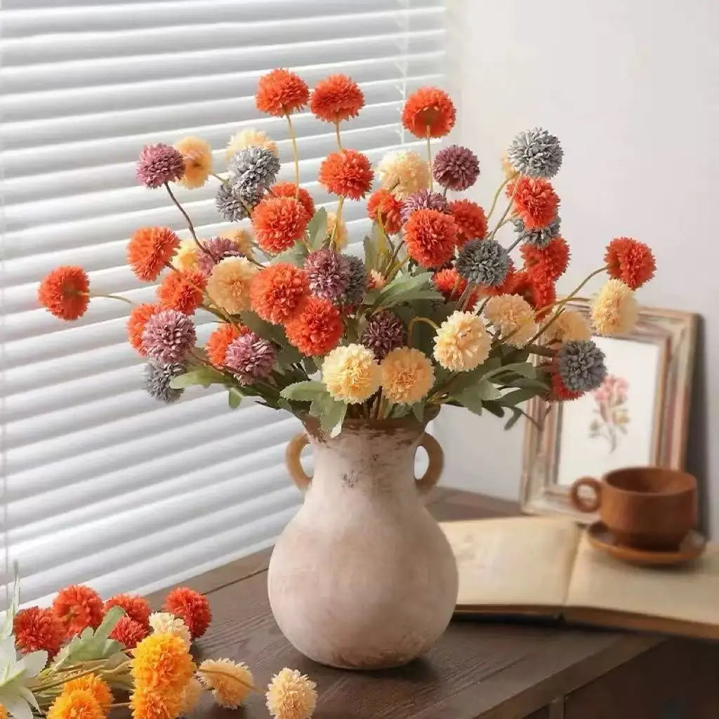 A bouquet of artificial orange, yellow, and purple chrysanthemum pompon flowers in a gray vase on a wooden surface.