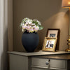 A dark gray vase filled with light pink and green flowers sits on a wooden dresser next to a framed photo.