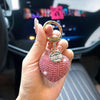 A hand holds a pink crystal heart keychain with a gold ring and a charm engraved with the name and date.