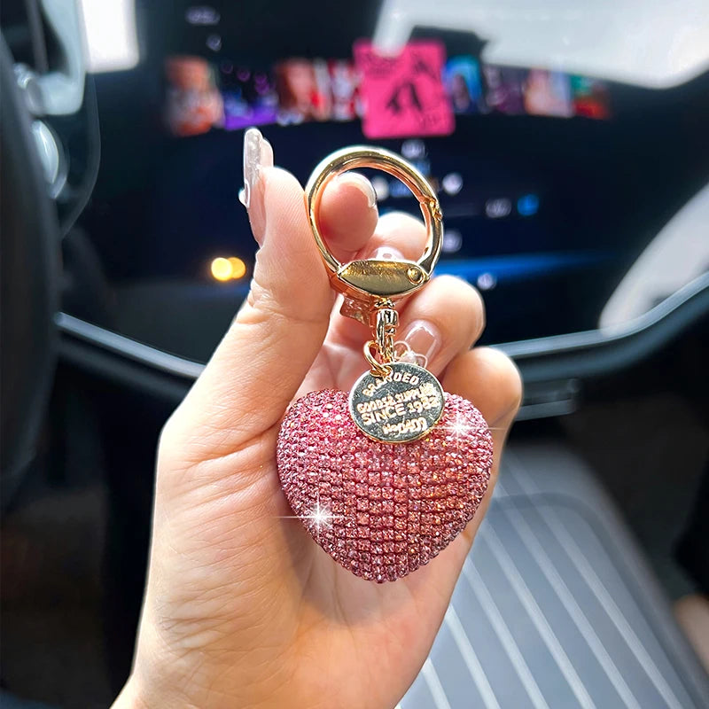 A hand holds a pink crystal heart keychain with a gold ring and a charm engraved with the name and date.