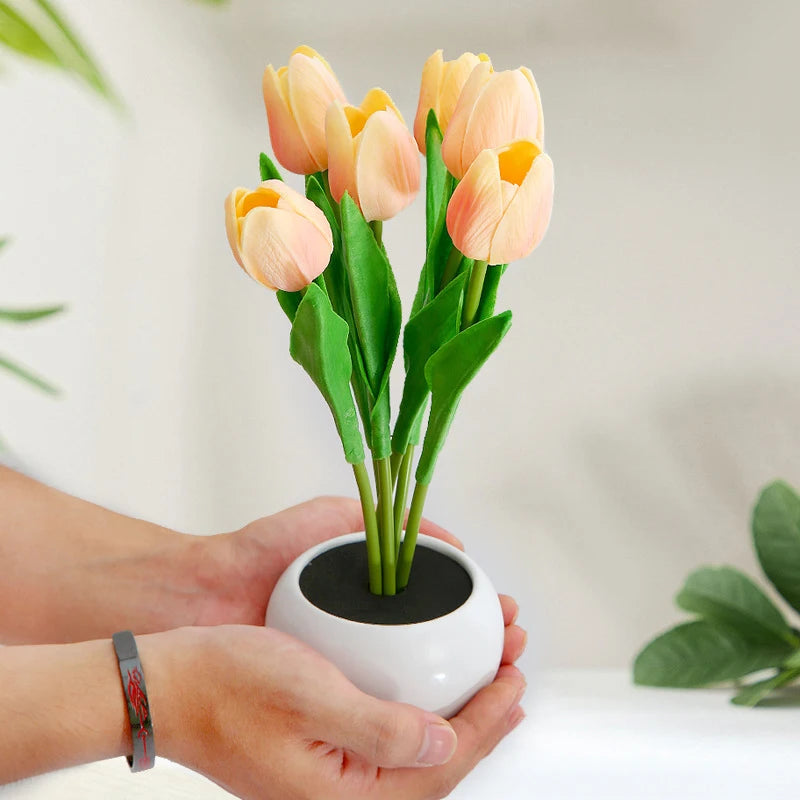 A hand holds a vase of pink tulips in a white ceramic pot, used as an LED night light.