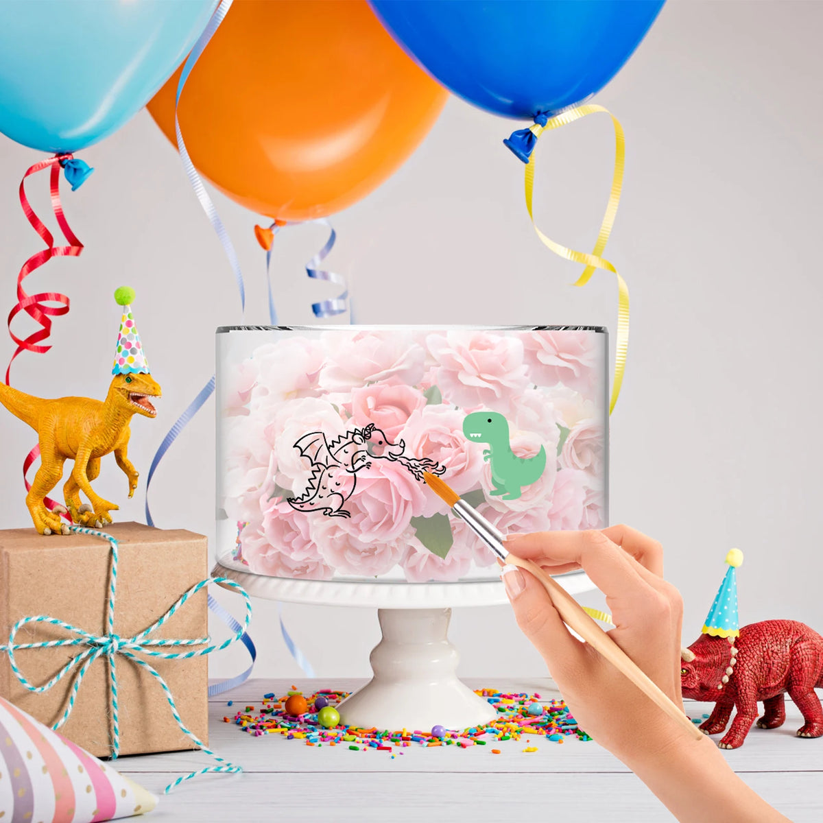 A hand paints a pink floral acrylic cake stand with dinosaur and bird designs on it, surrounded by colorful balloons and conf
