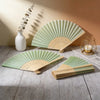A set of light green and natural wood folding fans displayed on a wooden surface with a white vase and gold wire stand.