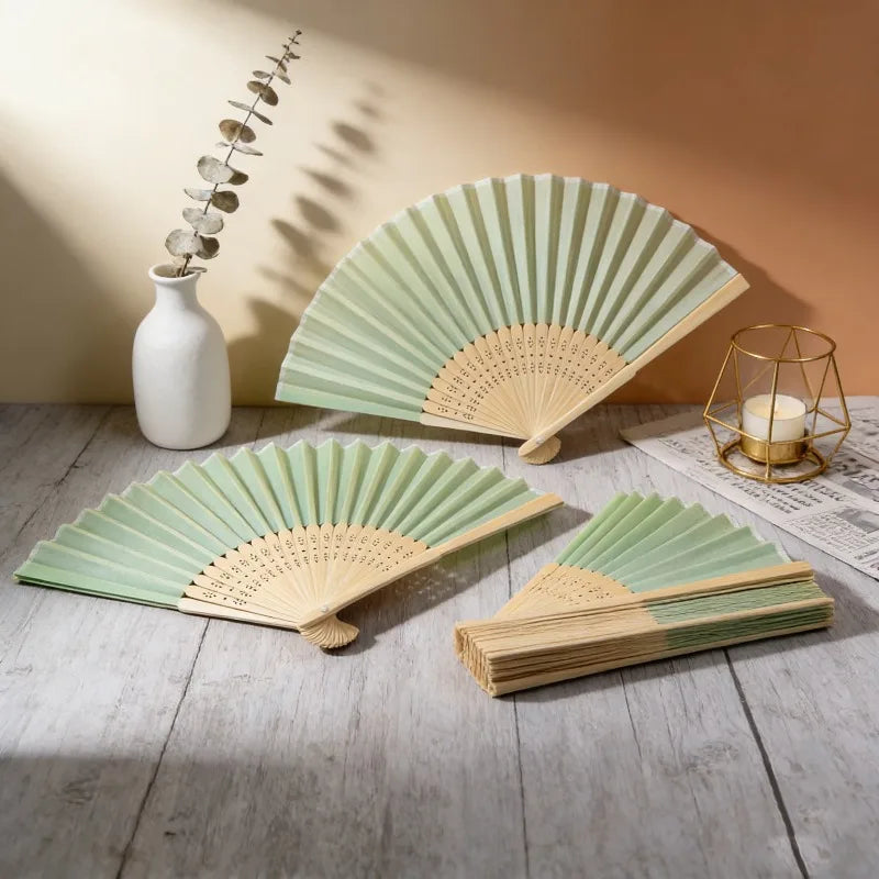 A set of light green and natural wood folding fans displayed on a wooden surface with a white vase and gold wire stand.