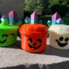 Three miniature pumpkin Halloween buckets in green, orange, and white sit on a gray surface.