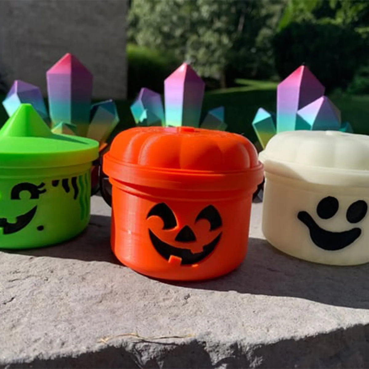 Three miniature pumpkin Halloween buckets in green, orange, and white sit on a gray surface.