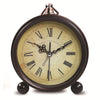 A vintage metal battery alarm clock with a round face and Roman numerals, set against a light brown background.