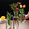 A clear glass hydroponic vase holds vibrant orange and pink flowers on a wooden table.