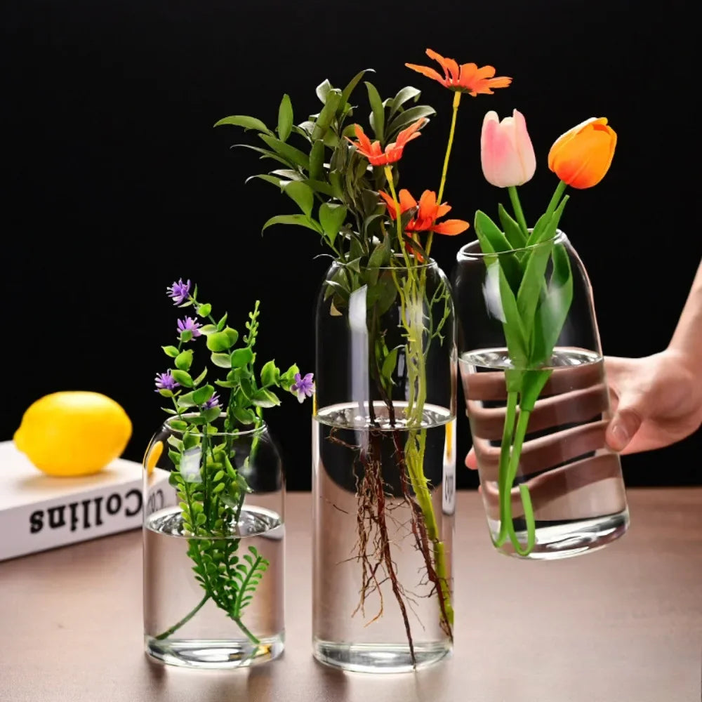 A clear glass hydroponic vase holds vibrant orange and pink flowers on a wooden table.