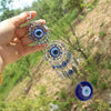 A hand holds a silver Turkish blue evil eye hamsa hand amulet with intricate details against a natural outdoor background.