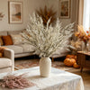 A vase filled with white artificial silk lover grass on a light-colored table in a living room setting.